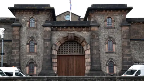 Exterior of HMP Wandsworth, a large brown brick building with dark wooden doors in the centre and windows, and columns, along the side. There are three white vans in shot along the outside. 