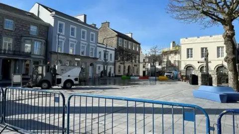 BBC Market Square, which is a large open space with grey paving stones on the ground and buildings around it. There are blue barriers in front  and a street sweeper cleaning the paving.