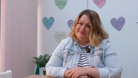 Alabaré Helen Brian has dark blonde shoulder length hair and is sat wearing a light blue denim jacket with her arms folded in front of her on a table. Behind her there is a plant and also some post-it notes on a wall displaying positive affirmations such as "it's ok to take things one step at a time".