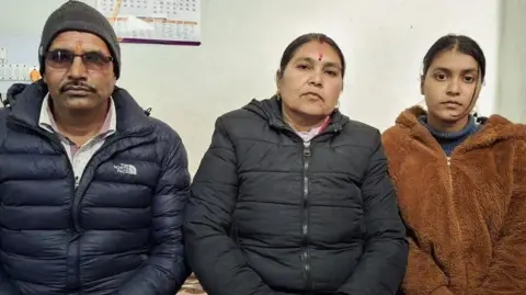 Mahananda, Padma and Puspa Joshi sit together in a row and look directly into the camera. There is a calendar on the wall behind them.