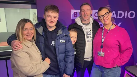 BBC Two women stand either side of two men with a boy in the centre. They are stood in a radio studio with a purple branded display board behind them reading 'BBC Radio Derby.' The woman on the left has a short blonde bob and a cream coat. The man to her right has his arm around her and is dressed in a navy hoodie, blue puffer jacket and has short fair hair. The boy in the centre is peaking his head out from behind the man. The woman on the far right has blonde hair tied back and is wearing a pink jumper, jeans, lanyard and glasses. The man to her left has short dark hair, a white hoodie, black open puffer jacket and dark trousers.