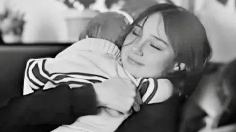 Family photo A black and white photo of a young woman with her eyes closed sitting on a sofa and hugging a small child. She has long dark hair and is smiling. She is wearing a dark long sleeve top. The child is wearing a light top with a dark horizontal stripe. 