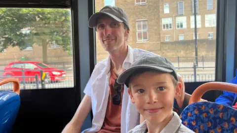 A man and his nine-year-old son sitting in blue and orange patterned seats on a bus. The man is wearing a grey baseball cap, white short-sleeved shirt and orange T-shirt. His son is wearing a matching cap and grey shirt.