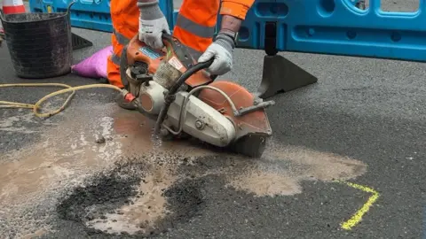 Chris McHugh/BBC A hand-held circular saw is operated by a man in orange hi-vis clothing. The saw is cutting into the tarmac around a pothole with water being produced from the saw