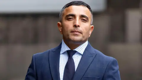 Abdul Nabizada arriving at court. He is wearing a blue suit over a light blue checked shirt and dark blue tie. He has dark hair on the top of his head which is greying at the sides.