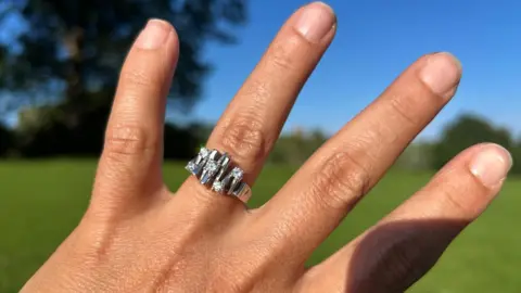 Nadya Giffen Close-up of a hand held against a blurred outdoor background of green grass, trees and a clear blue sky. A silver ring with multiple stones and geometric settings is worn on the ring finger. Bright sunlight highlights the ring and the texture of the skin, with the background softly out of focus to draw attention to the jewellery.