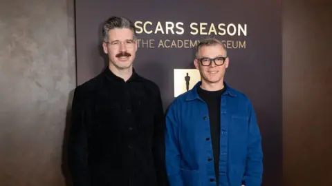 Getty Images Two men stand side by side in front of a backdrop featuring the words “OSCARS SEASON” and “THE ACADEMY MUSEUM.” The man on the left is wearing a dark, button‑up jacket, and he has hair grey, glasses and a moustache. The man on the right also has short grey hair, wears glasses, and is wearing a blue jacket over a dark shirt. The backdrop is in warm brown tones with an Oscars statuette icon displayed between the text elements.
