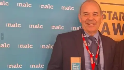 Image of a man in a black suit, grey shirt and navy tie who is smiling at the camera. He is standing in front of a large display board. Around his neck he has two lanyards, a red and a black lanyard - and he is holding an award.