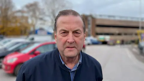 Phil Bialyk, leader of Exeter City council, wearing a blue check shirt and a navy blue jacket, in a car park.