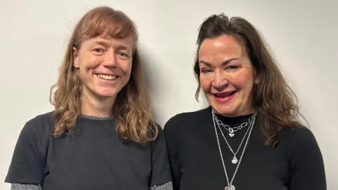 Sarah Bolger is wearing a grey shirt and has long, blonde hair and Justine Wortsman has three silver necklaces around her neck and is wearing a black shirt. She has long, brown hair.
