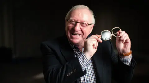 Mindhouse Productions Dennis Taylor, holding the pair of glasses he wore during the famous snooker match in 1985. He is wearing a new set of glasses and a black jacket of a check shirt.