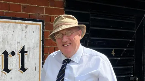 Matt Marvel/BBC Howard Watts is pictured outside the White Hart pub. He is wearing a cream coloured bucket hat and a light blue shirt with a navy blue and white tie. He has small glasses and is smiling at the camera.