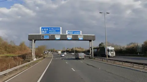 The M40 motorway, with cars and a lorry on it, showing six lanes of traffic, and an overhead sign that shows what junctions are coming up, with blue writing on. The sky is grey and dark. There is a lamp post to the left. 