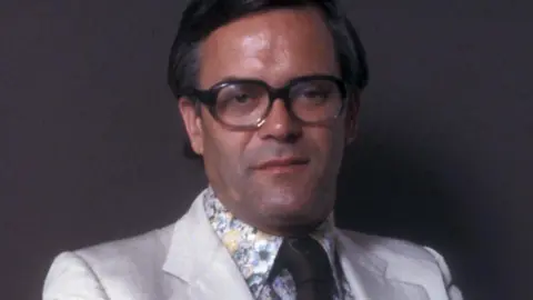 Sir Humphrey Burton pictured in a white suit, floral shirt and glasses