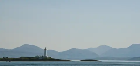 Lauren Clinch Landscape image with blue and grey tones of a lighthouse and calm waters with hills partially covered by fog in the far distance