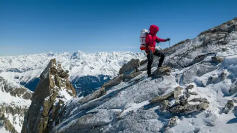 Jan Virt Fay Manners using ropes to reach the summit of the mountain. She is carrying equipment on her back and the sky is blue with snow covering the peaks of other mountains in the distance.