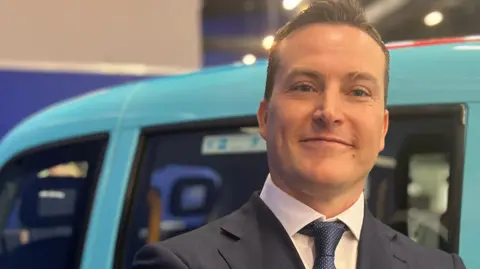 James McMurdock is in a navy suit and blue patterned tie, with what looks like a Hackney cab behind him, painted the sky blue colour of Reform UK.