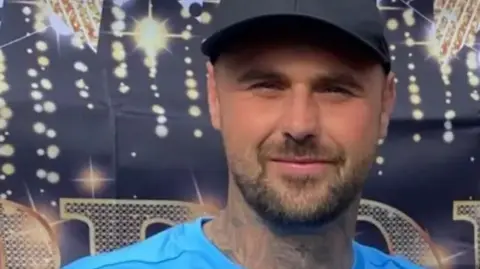 Supplied Tom Gaughan has a neatly trimmed, dark beard and tattoos on his neck. He is wearing a blue top and a North Face cap and is smiling into the camera.