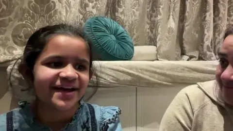 Fatima smiles off camera, her mother Lubna is sat next to her - cropped by the image. There are cushions behind her.