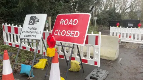 The picture shows road closed signs in red in front of white and red stripped fencing to block a road. 
