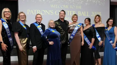 North West Anglia Hospitals’ Charity Seven women and one man wearing glamorous evening wear lined up with Craig Revel Horwood in the middle. He is wearing a gold spangled black jacket over a black shirt. They all have blue sashes on saying Table Hosts. They are all smiling broadly. Behind them is a screen saying Patron's Glitter Ball 2025.