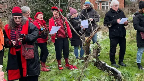 Irvine Road Community Orchard A group of people stand in a circle and sing together, some holding sheets with lyrics on. Some of the group are dressed in red and wear clogs as part of a Morris dancing troupe. They stand on a field with bare tree branches around them and a brick house behind.