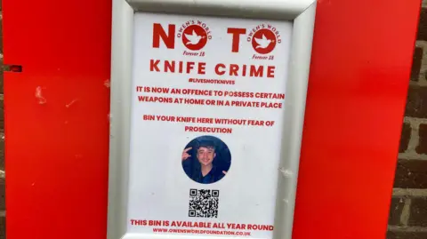 A notice attached to a bright red bin reading: "No to knife crime. It is now an offence to possess certain weapons at home or in a private place. Bin your knife here without fear of prosecution. This bin is available all year round."