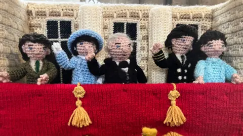 Tom Jackson/BBC A knitted scene showing members of the royal family and Winston Churchill waving and standing on Buckingham Palace balcony in 1945. From the left, Princess Elizabeth is in the khaki uniform of the ATS, Queen Elizabeth is in bright blue, Winston Churchill is in a black suit, King George VI is wearing a naval uniform and Princess Margaret is wearing light turquoise. The balcony is draped in red and has yellow tassels.