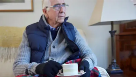 An elderly man sits on a white sofa. He is looking out a window out of picture, wearing a grey cardigan, a body warmer, jumper, shirt and black gloves. He has white hair and wearing glasses. To the right of where he is sitting is wooden lamp with a white shade. He is holding a teacup and saucer. 