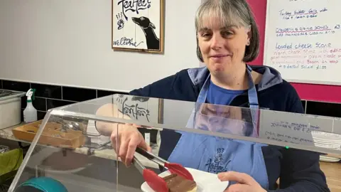 TeaHee Cafe A woman wearing a blue apron, standing behind the counter in a coffee shop smiling at the camera. She is holding a plate with a millionaire's shortbread on it, held in place with a pair of tongs. A sign behind her says "TeaHee".