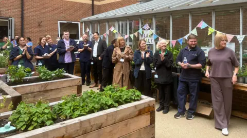 Jessica Bayley/BBC A group of people all stood in front of a greenhouse with bunting on it, they are clapping and smiling. Around them are raised flower beds.