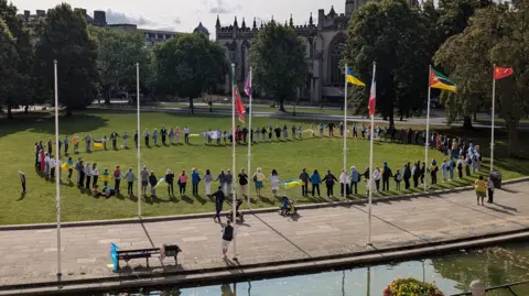 Antonina Grebeniuk A circle of people standing on the grass of College Green holding hands. The photo was taken from a slightly elevated position. Several flags can be seen, including the Ukrainian flag.