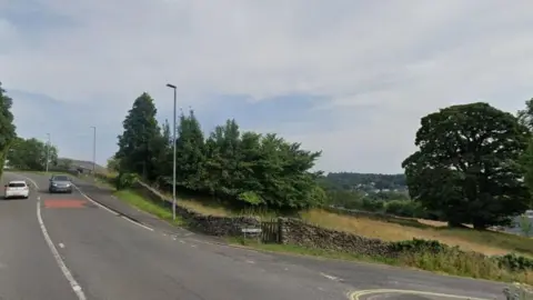 Google The current junction from the A591 and Thwaites Lane into the Orrest Head Farm site. There are trees and fields at the corners.