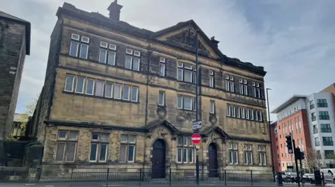 The exterior of the former Gibson Street Baths building. It is three-storeys high and built from stone with several windows running along the front. Two dark wooden doors stand as separate entrances for men and women. 