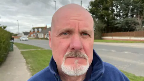 John Devine/BBC Martin Reynolds, 62, has a bald head with a neat grey beard and moustache, he is wearing a blue fleece top with the collar up around his neck, behind him is a roadway with trees and houses visible and a white  moving car on the left.