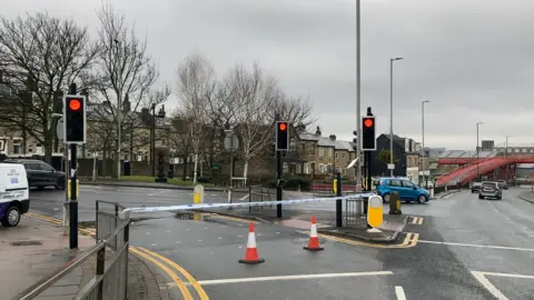 Police tape across a traffic light crossing with cars going past. There are cones in front of the police tape.