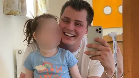 Family handout Joshua Hardman with brown hair wearing a white t-shirt and jeans crouches  down to sit behind his young smiling daughter wearing a light blue t-shirt sitting on a low stool. Joshua holds up a white mobile phone.