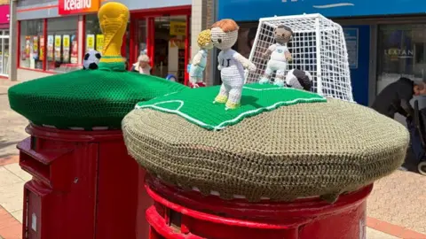 YaY Yarn Bombers Two red post boxes on a high street topped with knitted creations. On the right, three male footballers in white shirts stand in front of a goal, one red-headed, one blond, one dark. A large knitted football is in the goal. On the left, the yellow World Cup trophy sits on a green knitted base with a black and white football next to it.