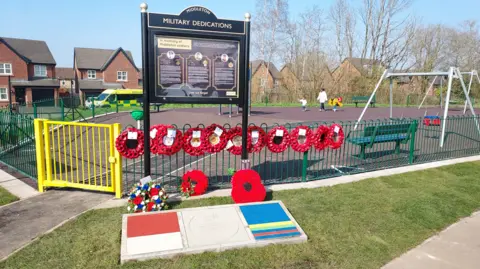 A black and gold sign says 'Middleton military dedications' and 'In memory of Middleton soldiers', with photos and dedications to the three soldiers. Multiple poppy wreaths have been laid on the green railings behind the sign and a stone memorial in honour of Corporal Joel Halliwell. A children's playground and some houses can be seen behind the sign.