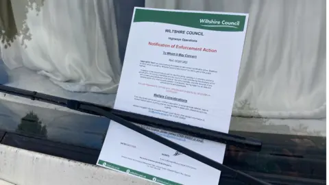 A notice tucked under a windshield viper. It is a letter from the Wiltshire Council, notifying the vehicle owner of 'Enforcement Action'.
