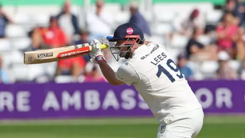 Left-handed batter Alex Lees, wearing "LEES 19" on the back of his white shirt, playing a shot through the offside for Durham, with a helmet on and a high bat.