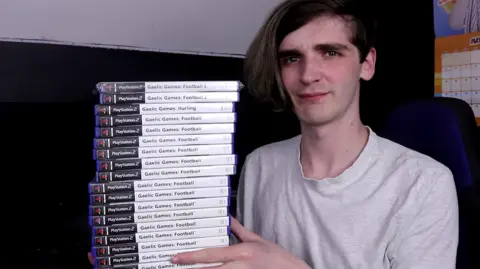 Matt Murphy A man looks a the camera. He is wearing a grey t-shirt and has dark brown hair, swept to the right-hand side. He is sitting on a sofa in what appears to a living room. He is holding 18 boxed PlayStation 2 copies of the three titles from the Gaelic Games video game series - Gaelic Games: Football, Gaelic Games: Football 2 and Gaelic Games: Hurling