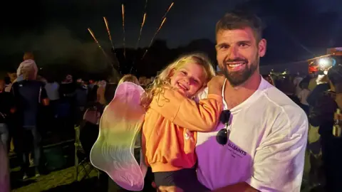 Alan Johnstone Alan Johnstone smiling, wearing a white t-shirt and holding his daughter Sienna with long blonde hair and also smiling, wearing an orange jumper with fireworks in the background