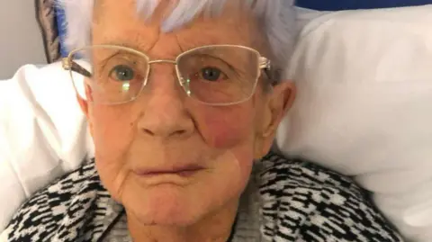 Maureen Harman, 88, who has white hair and golden rimmed glasses, looks into the camera with a serious expression while sitting up on a hospital trolley.