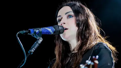 Amy Macdonald performs onstage, singing into a microphone. She has long dark hair and is playing a guitar