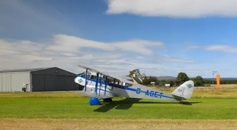 Walter Dalgleish A blue-and-white vintage biplane sits on a grassy airfield beside a large hangar. The aircraft is parked under a bright blue sky with scattered clouds, with countryside and buildings visible in the background.