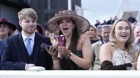 PA Media A woman in a green dress and purple hat cheers while holding a mobile phone as a man in a blue suit and tie and a woman in a white and black patterned dress stand either side of her