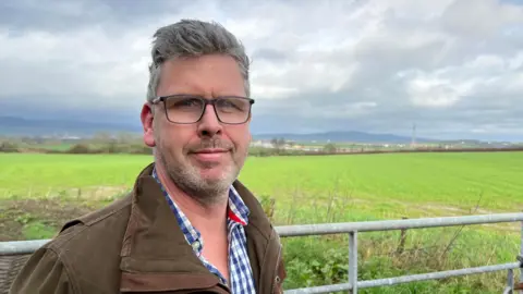 Councillor Merrett stood in front of field with small hills in the background. He is wearing a checked shirt, brown coat and glasses.
