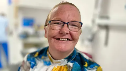 Kathryn Finlay is wearing a blue and gold high-neck satin top and is smiling at the camera. She has some teeth missing. She has fair reddish hair pulled back into a ponytail and is wearing black-rimmed glasses. 