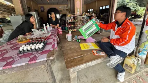 Two women in a shop are talking to Chutiphong Pipoppinyo who is sitting on a bare wooden table and showing them a mockup of a ballot paper. Pipoppinyo is wearing blue jeans, white sneakers and an orange and white jacket. 